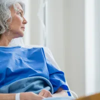 Kuvassa on harmaahiuksinen nainen potilasvaatteissa. Hän istuu potilasvuoteella ja katsoo ulos ikkunasta.
