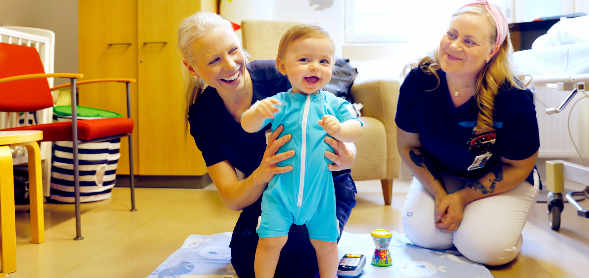 Fysioterapeutti Taru Palsa pitelee pystyssä yhdenksänkuista Rafael-vauvaa. Tutkimushoitaja Jonna Salama istuu vieressä.