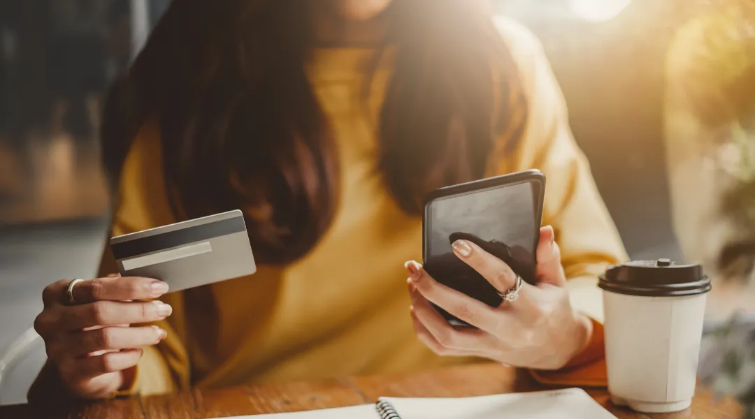 Nainen, jolla on ruskeat pitkät hiukset ja keltainen paita pitelee toisessa kädessä luottokorttia ja toisessa kädessä matkapuhelinta.