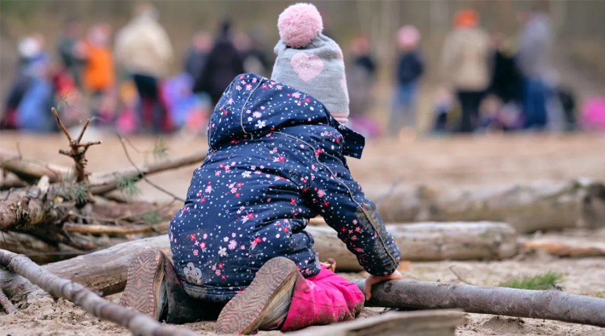 Selin olevan pikkulapsi leikkii maassa, taustalla on kauempana aikuisia.