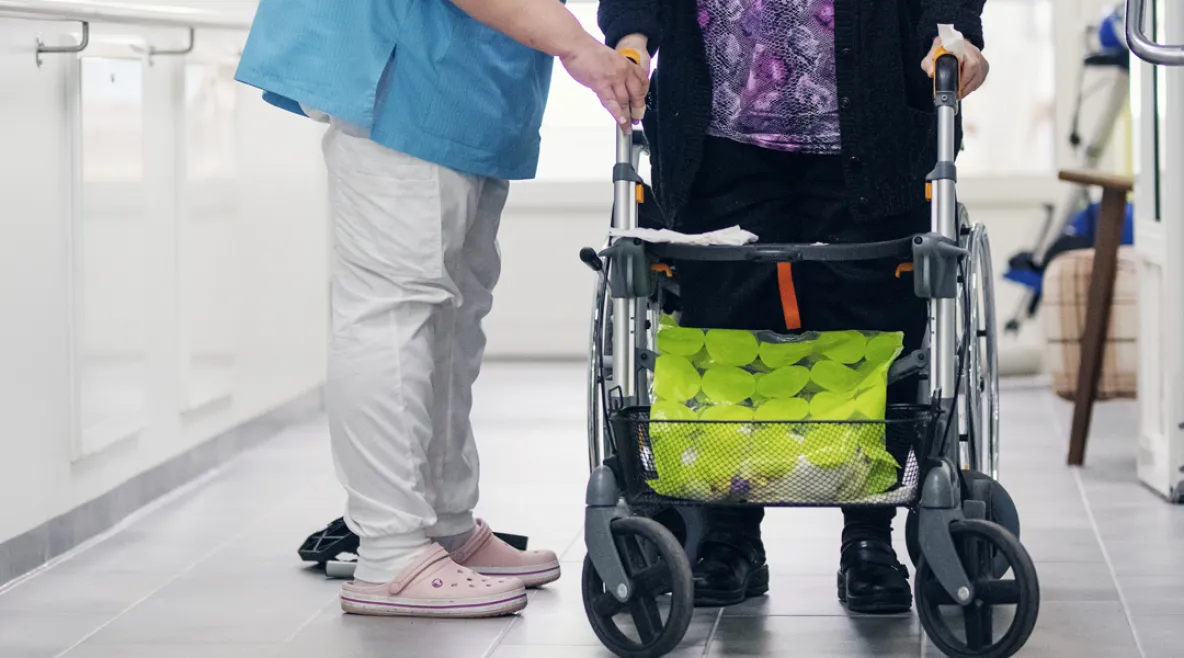Hoitaja auttaa rollaattorin kanssa liikkuvaa vanhusta laitosoloissa.