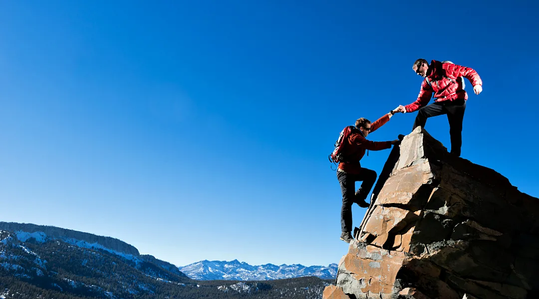 Kuvassa toinen vuorikiipeilijä auttaa toista nousemaan terävän vuorenhuipun päälle.