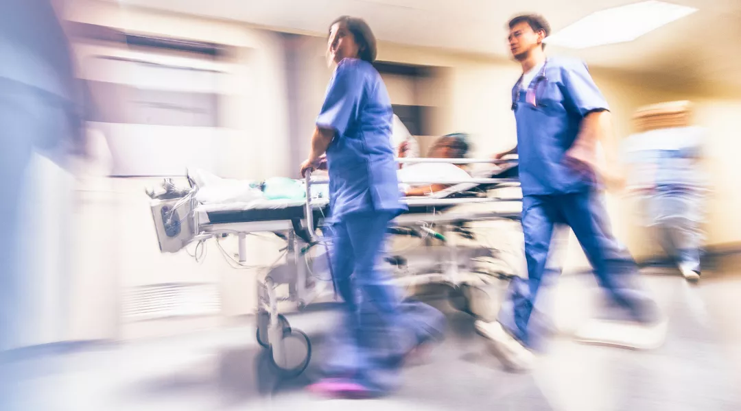 Kuvassa kaksi sairaanhoitajaa sinisissä työasuissa kiidättää potilasta potilasvuoteella sairaalan käytävällä oikealta vasemmalle.