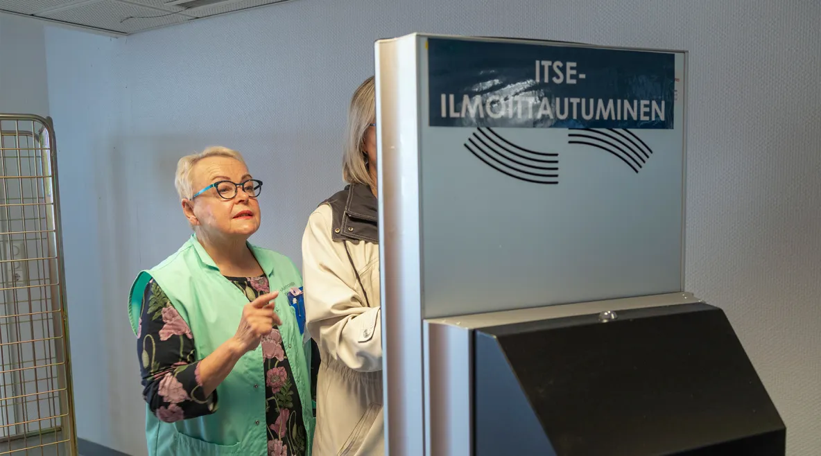 Itseilmoittautumisautomaatilla tuntematon asiakas, jota vaaleanvihreään vapaaehtoisten liiviin pukeutunut Thea Nurmi neuvoo.