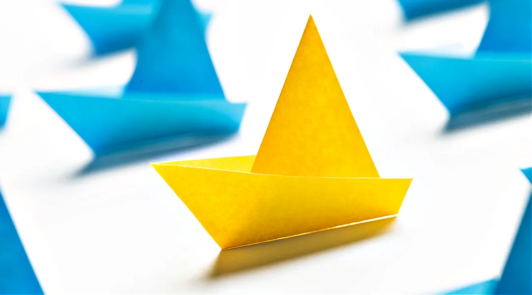 Kuvassa on keltainen paperista taiteltu origamivene ja taustalla sinisiä origamiveneitä.