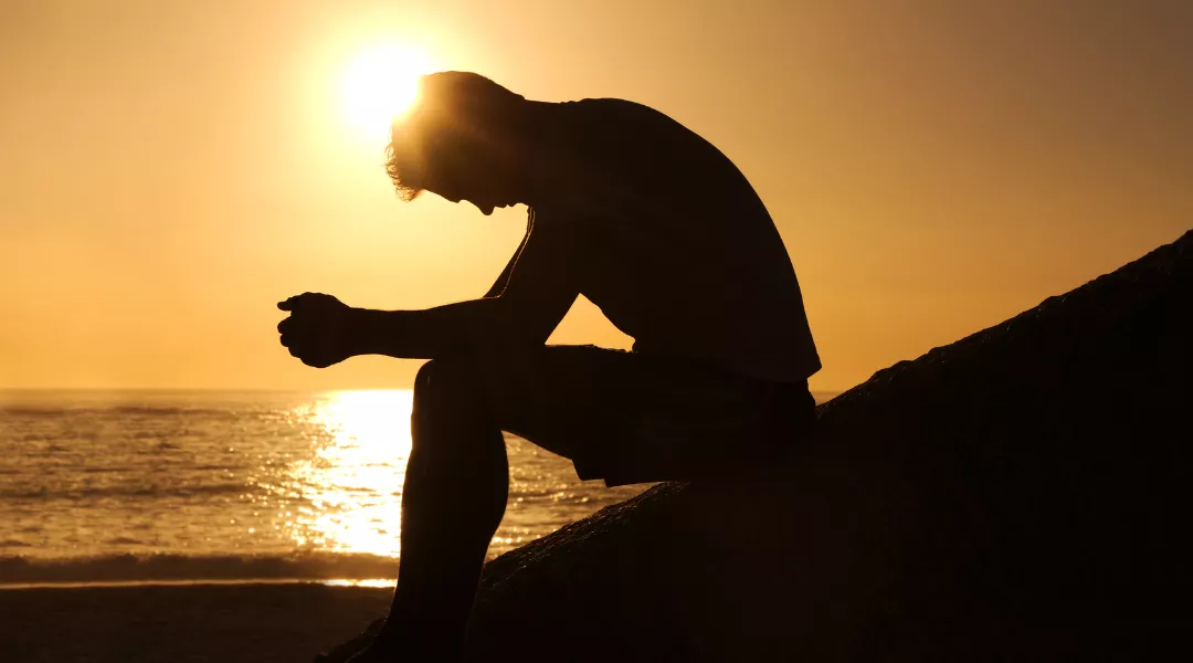 Kuvassa on miehen siluetti sivusta, joka istuu merenrannalla pääpainuksissa ja takaa paistaa aurinko.