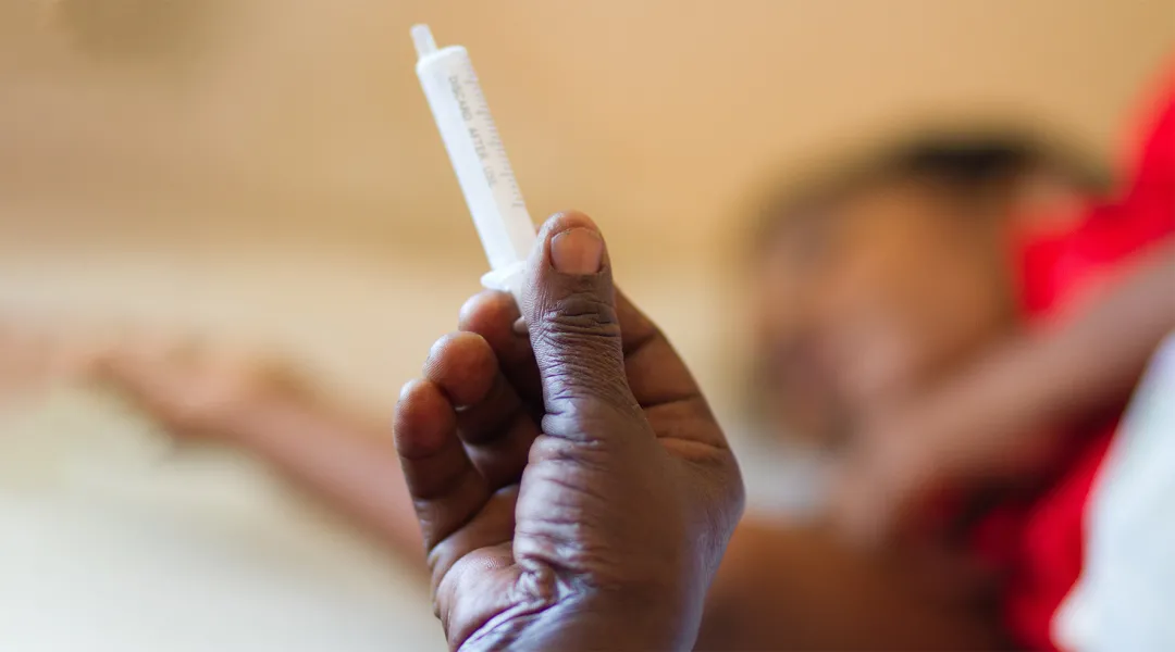 Sambialainen hoitaja pitää kädessä ruiskua. Potilas näkyy taustalla. 