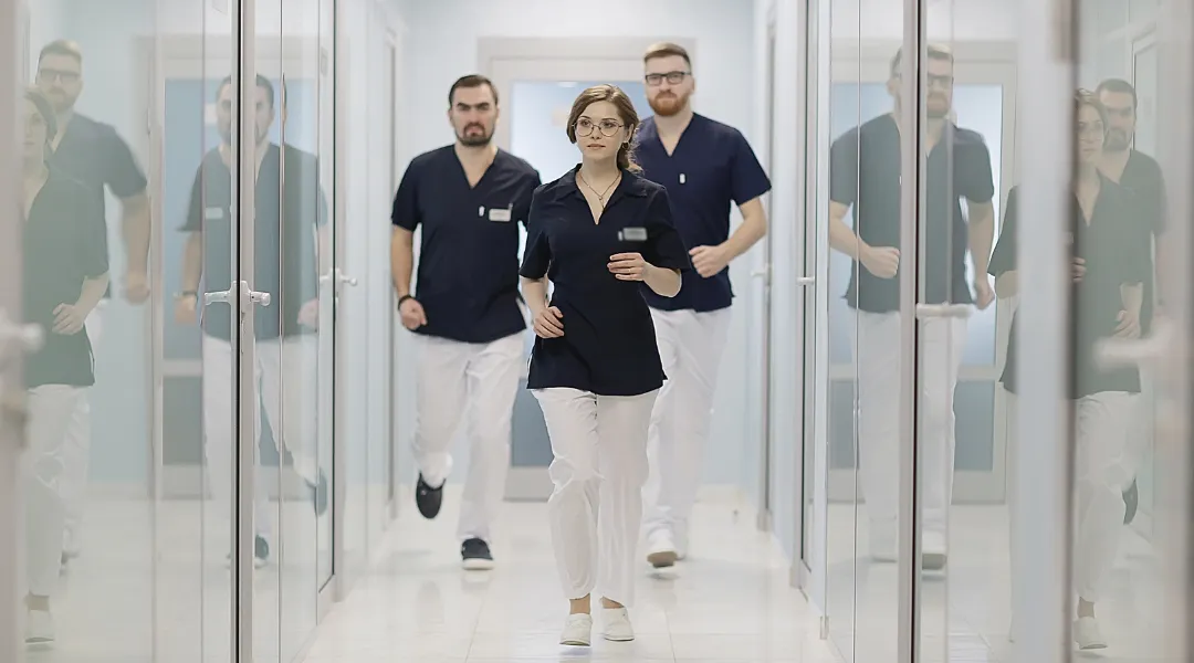 Kolme sairaanhoitajaa jouksee käytävällä sinivalkoisissa työasuissa.