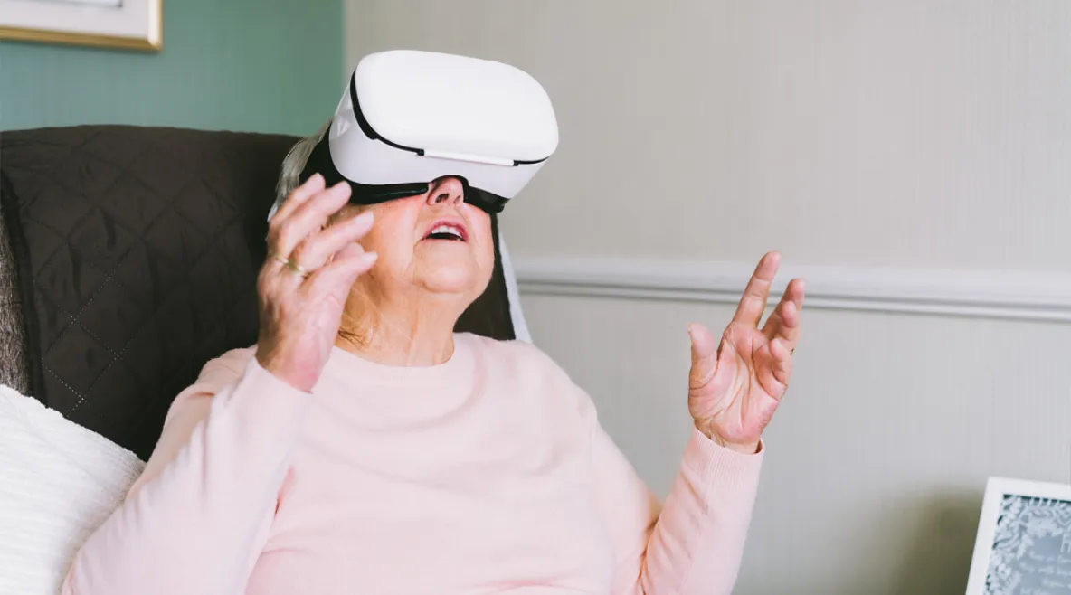 Vanhempi nainen istuu tuolissa vr-lasit päässään ja kurottelee käsillä ylöspäin.