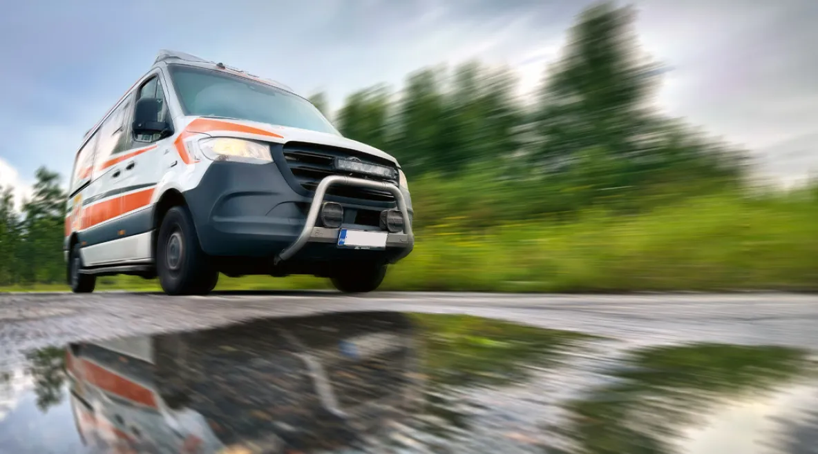 Ambulanssi ajaa kovaa vauhtia asfalttitietä, jonka päällä vesilammikko.