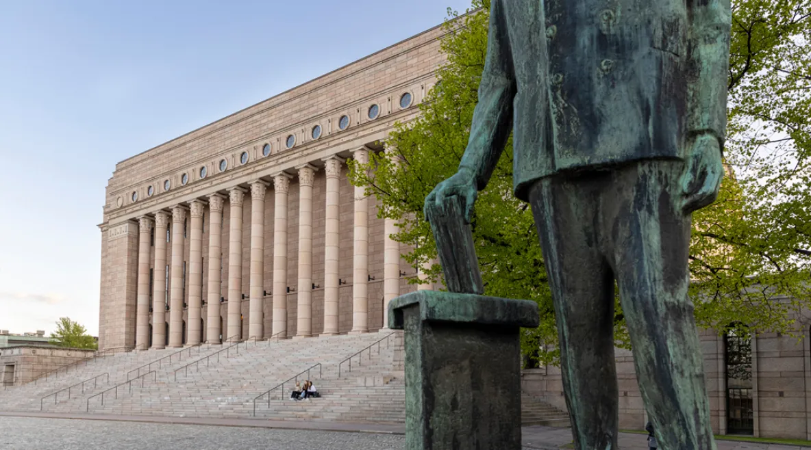 Eduskuntatalo Helsingissä kevätauringossa. Etualalla näkyy työväenjohtaja Yrjö Mäkelinin patsas, jonka hahmo pudottaa paperin vaaliuurnaan.