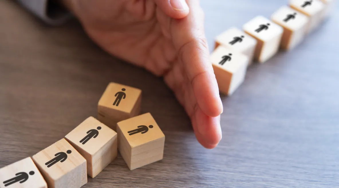 Laboratorioyhtiö Fimlab on aloittanut muutosneuvottelut, jotka voivat johtaa 30 henkilötyövuoden vähentämiseen. Käsi estää puisista palikoista muodostetun ihmisjonon etenemistä, yksi palikka on kaatunut muiden edessä.