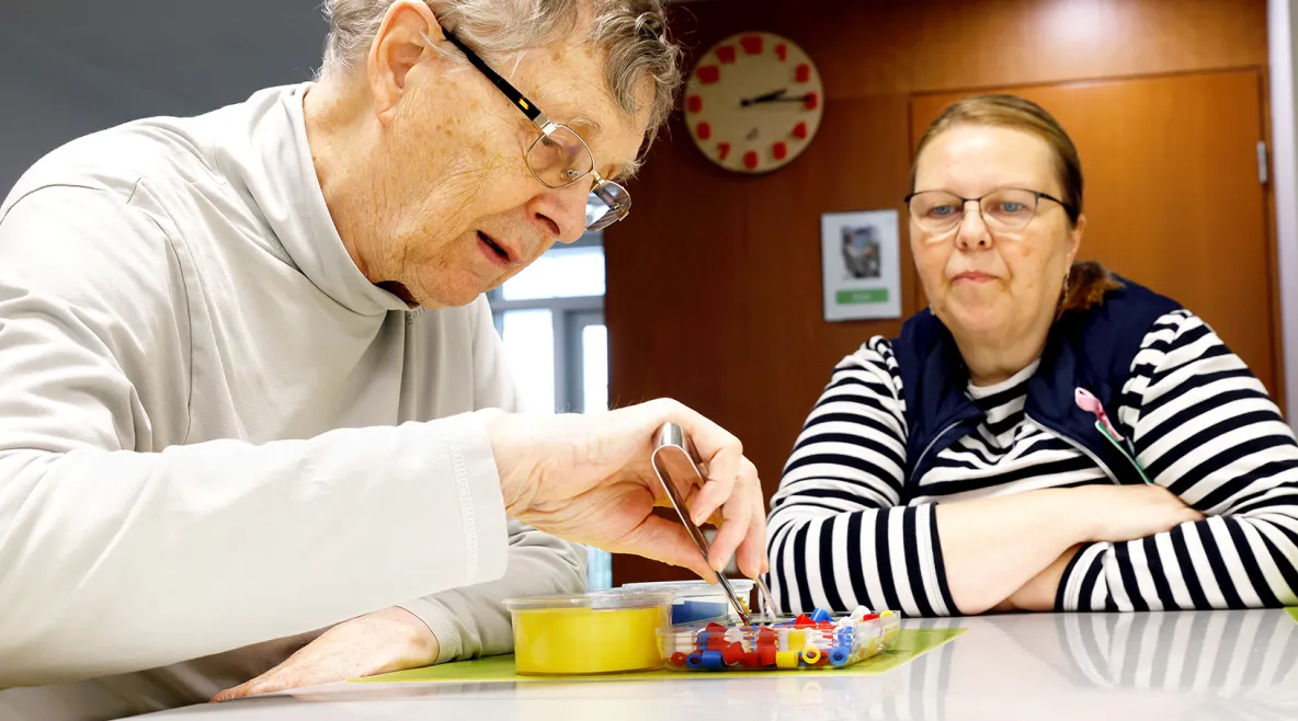 Markku Närhistä auttamassa lähihoitaja Marianne Vierelä. Markku lajittelee pinseteillä pieniä muoviesineitä värin mukaisiin rasioihin.