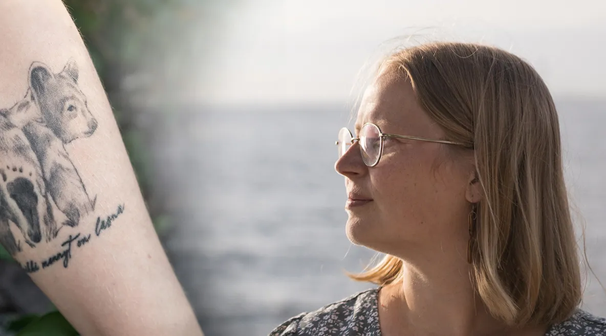 Kuva kädestä, jossa karhutatuointi. Oikealla Sairaanhoitaja Mirva Virtanen katsoo horisonttiin taustalla meri.