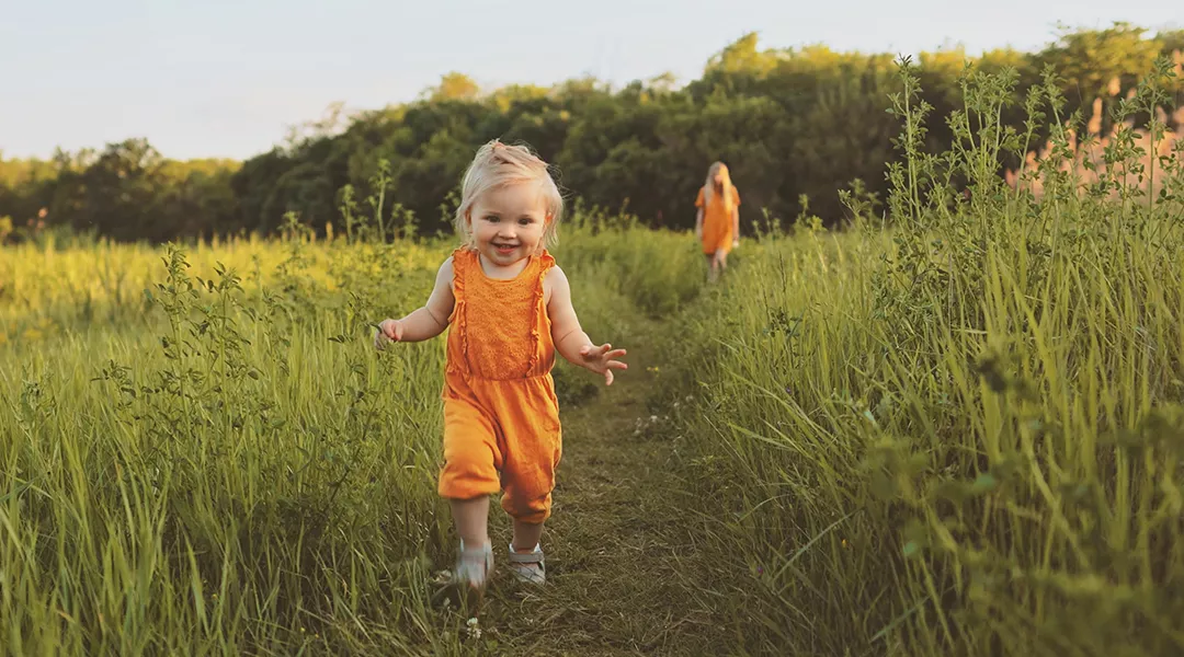Lapsi kesäisellä niityllä, äiti kävelee kauempana taustalla.