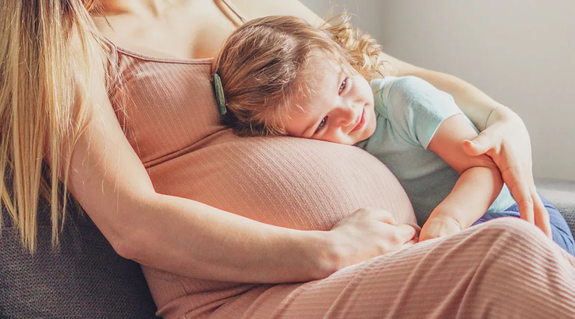 Raskaana oleva nainen, johon isompi lapsi nojaa.