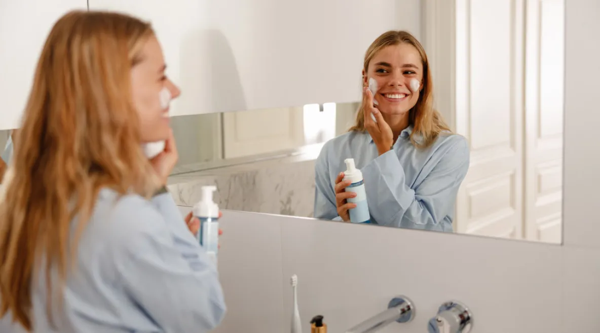 Nainen levittää voidetta kasvoilleen kylpyhuoneessa ja katsoo itseään samalla peilistä hymyillen.