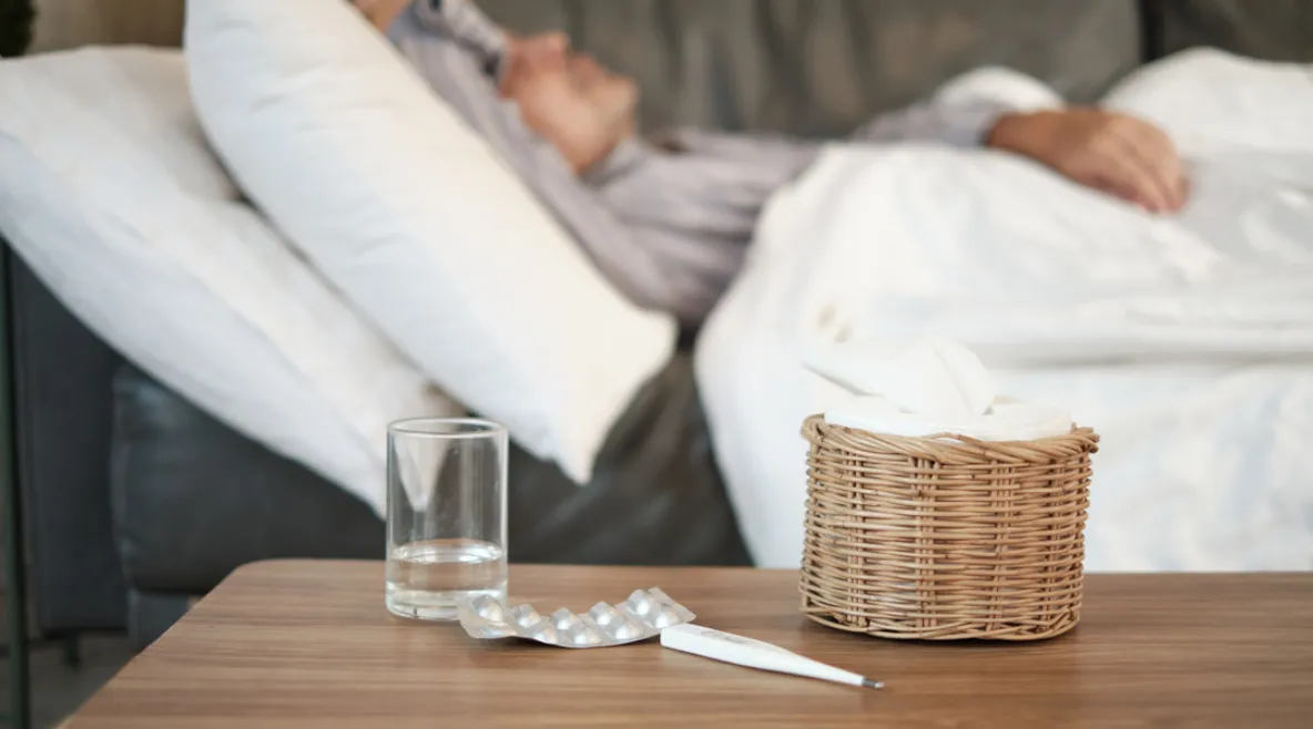 Influenssa jyllää, mutta rokote suojaa vakavalta taudilta. Sairastunut mies makaa sohvalla, pöydällä vesilasi, särkylääkkeitä ja kuumemittari.