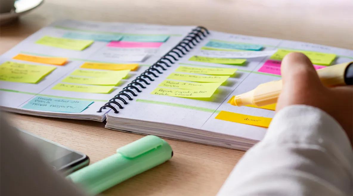 Henkilö kirjoittaa keltaisella korostuskynällä muistivihkoon, joka on täytetty värikkäillä post-it-lapuilla ja tehtävälistoilla. Vihko on avoinna pöydällä, jonka ääressä näkyy myös toinen korostuskynä ja älypuhelin.
