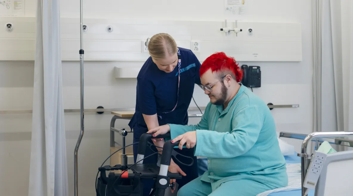 Hoitaja auttamassa potilasta nousemaan kävelytuen avulla ylös sairaalasängystä.