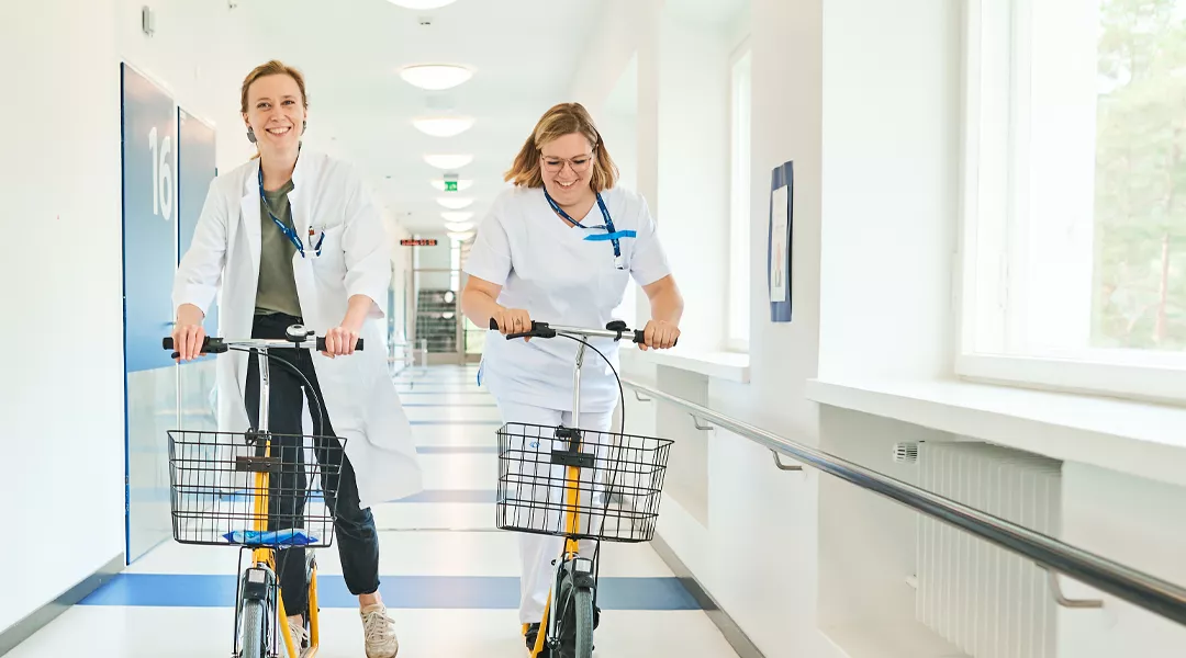 Kaksi hoitajaa potkupyöräilee sairaalan käytävällä.