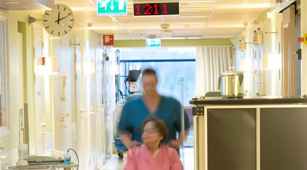 Hoitaja työntää potilasta pyörätuolissa laitoksen käytävällä.