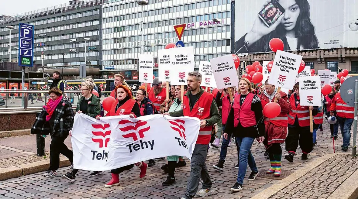  Tehyn jäseniä marssii kadulla Helsingissä punaisiin liiveihin pukeutuneina ja kantaen banderollia, jossa lukee &quot;Tehy&quot;. Useilla on kylttejä, joissa vaaditaan parempaa palkkaa ja arvostusta hoitajille. Taustalla näkyy Elielinaukio, Yliopiston Apteekki, mainostauluja ja Helsingin keskustan rakennuksia.