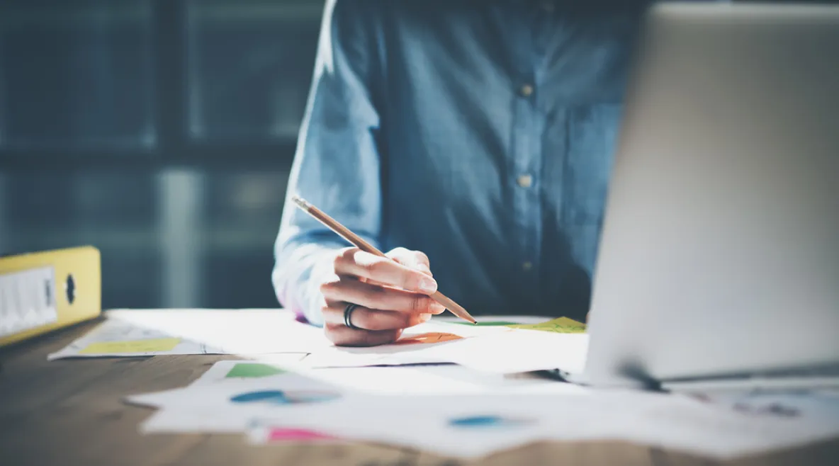 Henkilö laatimassa tietokoneella suunnitelmaa ja hahmottelee samalla kynällä paperille.