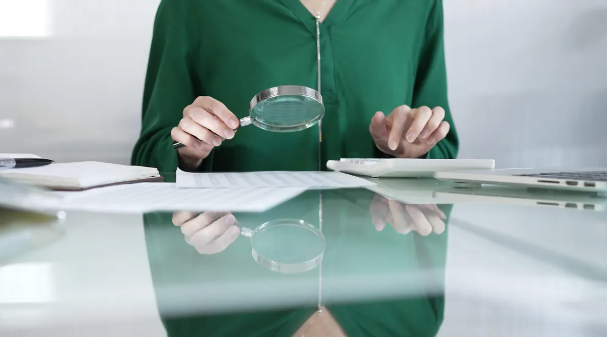 Ihminen istuu pöydän ääressä ja tutkii suurennuslasilla papereita. 