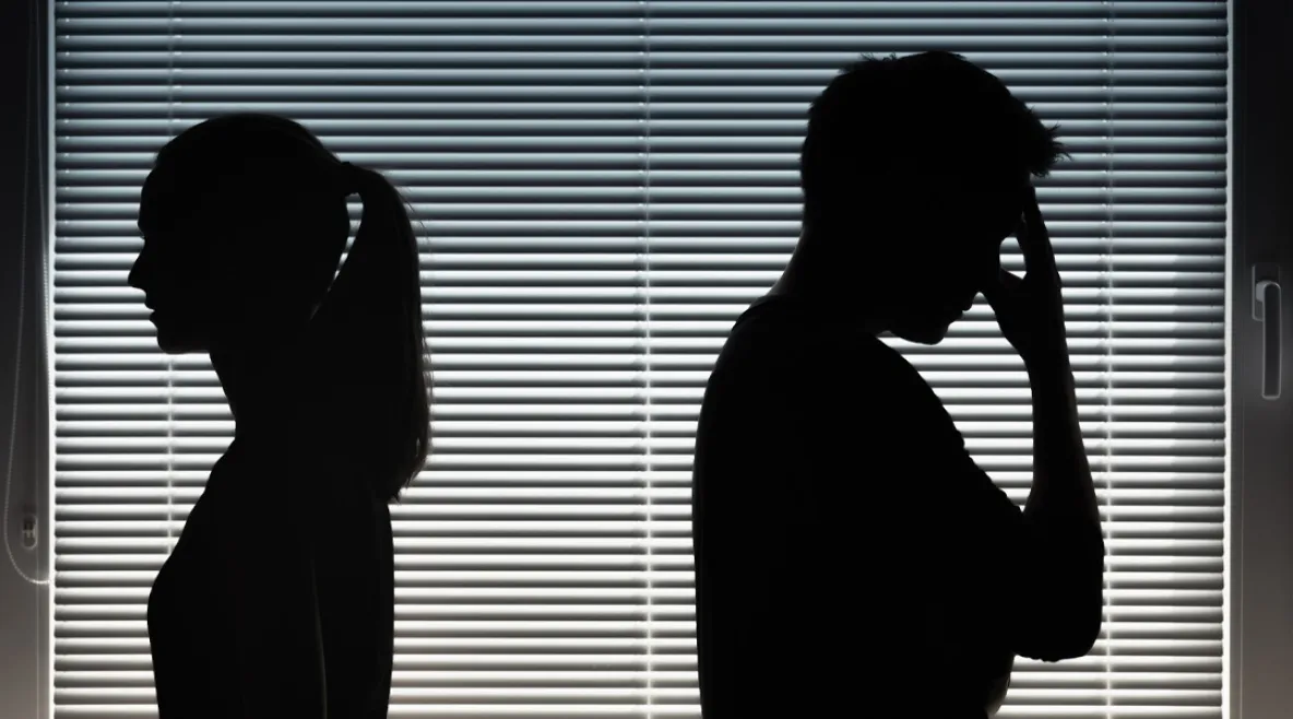 Miehen ja naisen tummat varjosiluetit sälekaihtimilla peitettyä ikkunaa vasten selin toisiinsa.