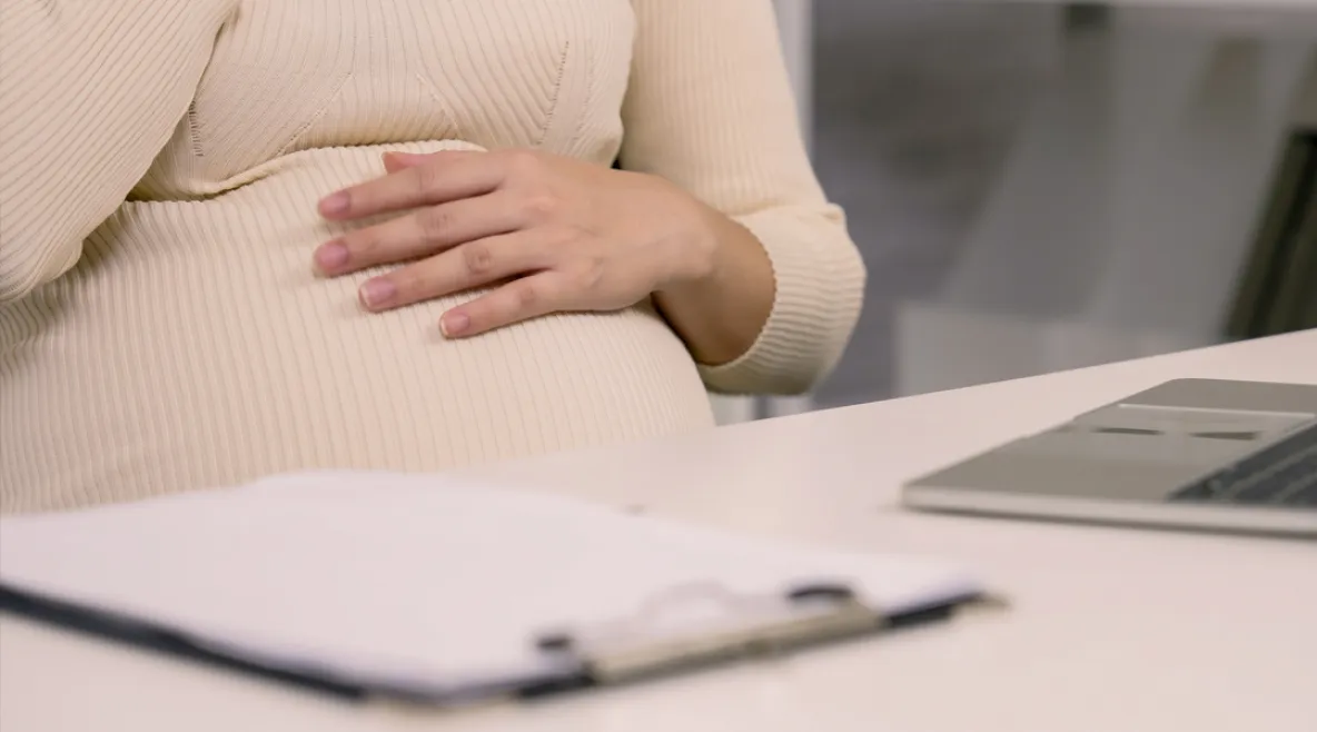 Raskaana oleva nainen pöydän ääressä. Hän pitää kättä pyöristyneen vatsanpäällä. Pöydällä lehtiö ja kannettava tietokone.