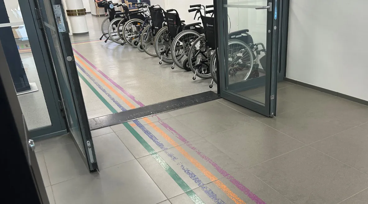 Kuva sairaalan aulatilasta, jossa eri väriset viivat johdattavat kävijän oikeaan paikkaan. Taustalla seinustalla pyörätuoleja.