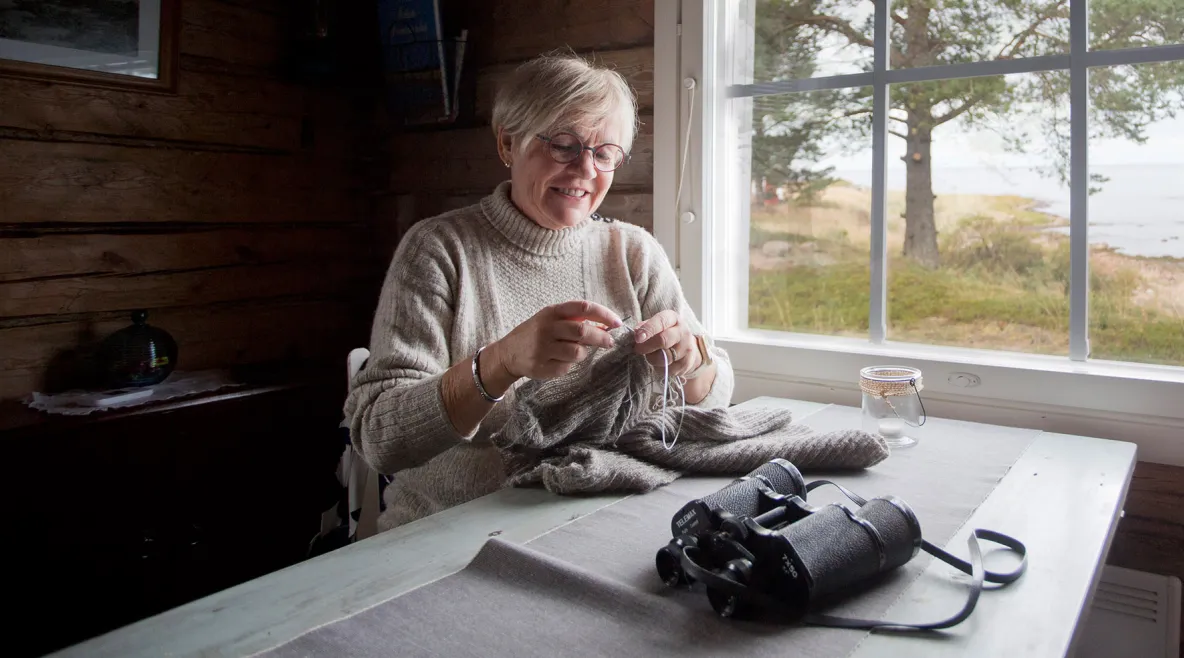 Tuovi Anttila neuloo mökillä pöydän ääressä ikkunan edessä.