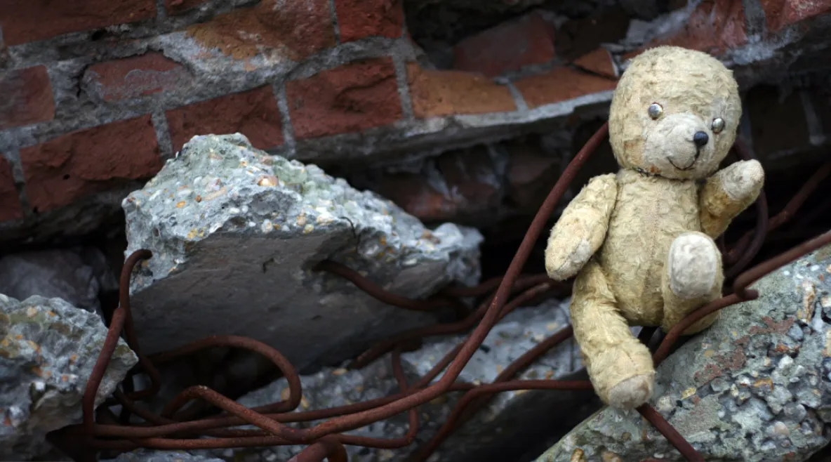 Kulunut ja likainen nallekarhu istuu sortuneen tiiliseinän ja betonilohkareiden keskellä, ruosteisten raudoitusten lomassa.