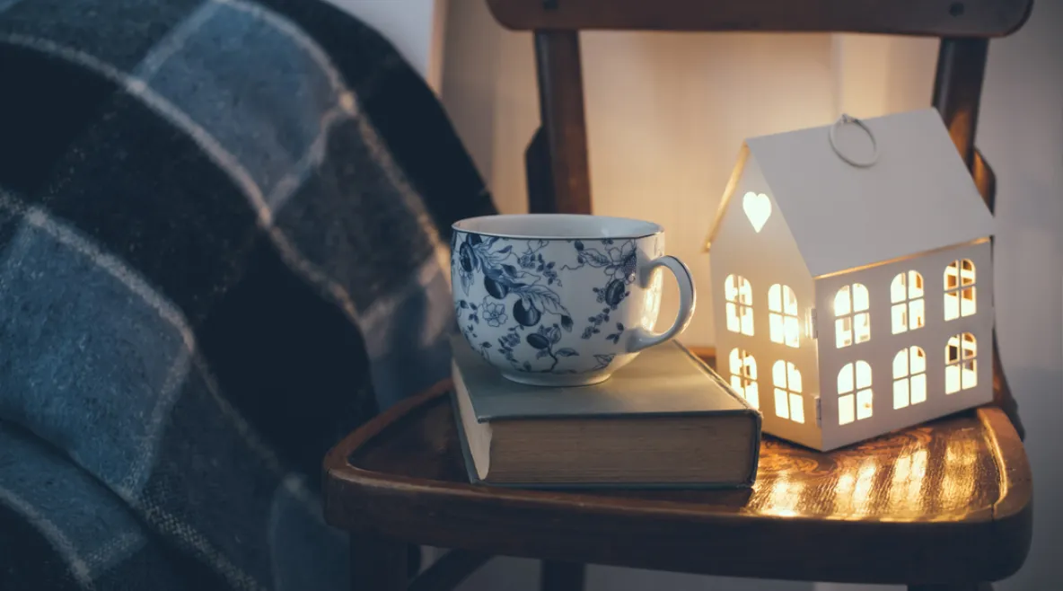 Tuolin päällä tunnelmallinen talolyhty, kirja ja kahvikuppi. Vieressä sänky.