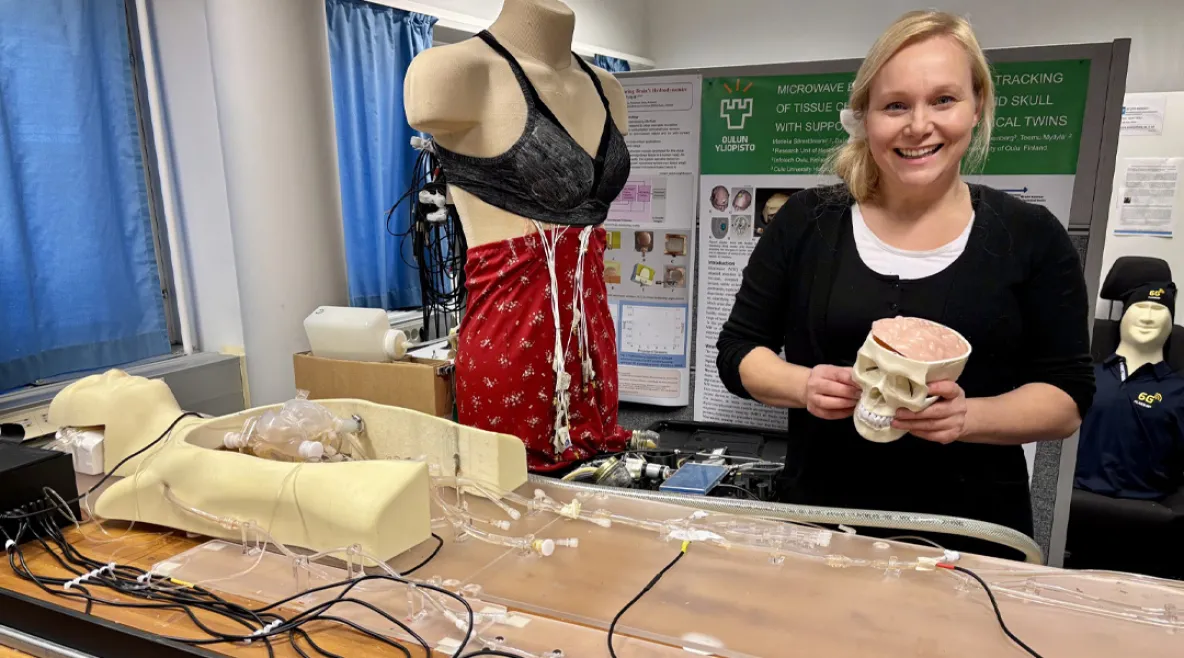 Yliopistotutkija Mariella Särestöniemi Human Body Twin -laboratoriossa, jossa kehitetään teknologiaa, joka mahdollistaa terveydentilan seurannan ja arvioinnin kannettavilla ja päälle puettavilla mittalaitteilla. Kädessään hänellä on aivomalli, jota käytetään signaalin etenemisen tutkimiseen aivokudoksessa, ja taustalla nukella on rintasyövän tunnistukseen kehitettävät monitorointiliivit. 