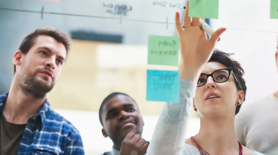 Työtiimi tekee suunnittelutyötä yhdessä kiinnittäen post it -lappuja lasiseinälle.