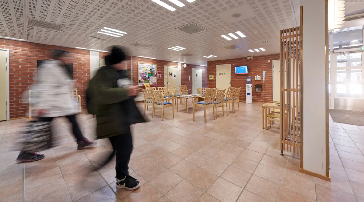 Terveyskeskuksen odotustila. Muutama ihminen epätarkkana kuvan etualalla kulkemassa huoneen läpi.