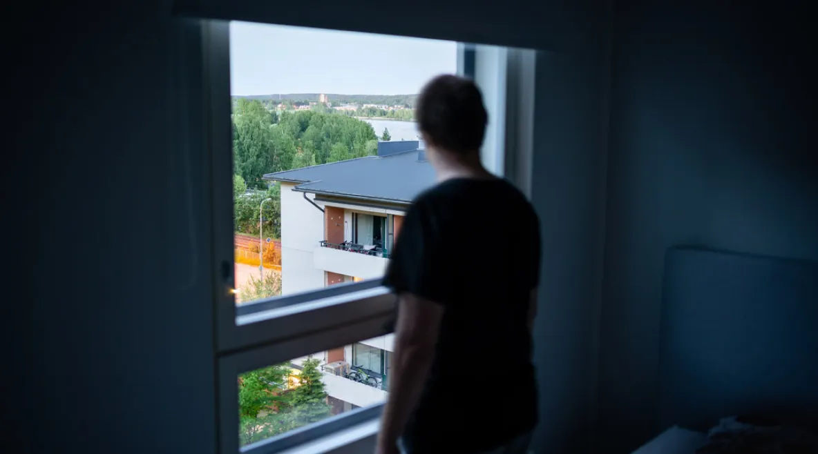 Ihminen katselee hämärässä huoneessa ikkunasta ulos lähiömaisemaa.