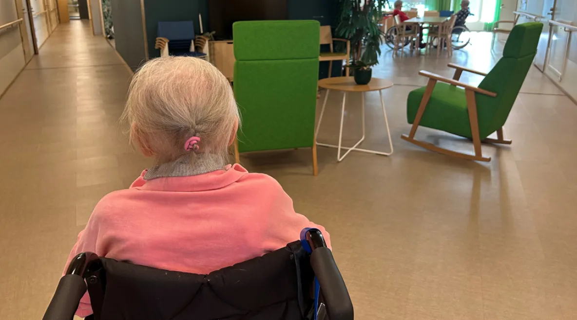Vanhus istuu pyörätuolissa selin kameraan hoivakodin aulatilassa.