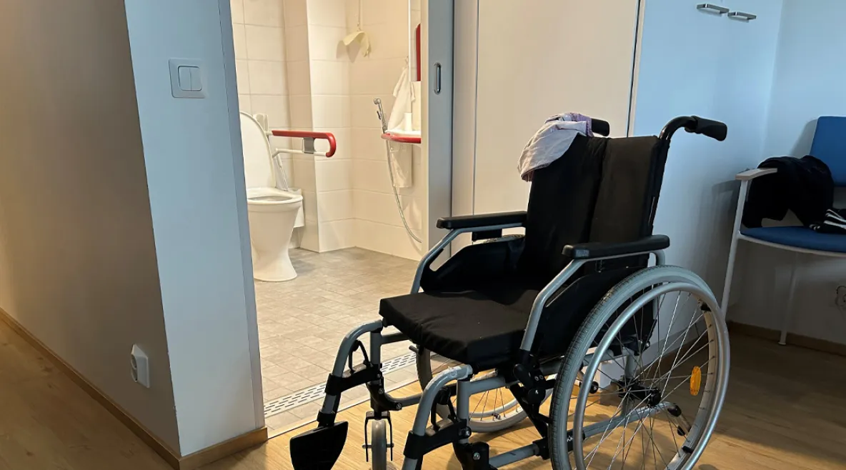 Pyörätuoli avoimen kylpyhuoneen edessä. Kylpyhuoneessa on tukikaiteet ja wc-istuin.