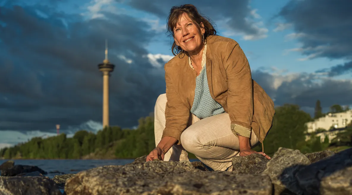 Ulla Ollikkala kyykyssä rantakivikossa, taustalla Tampereen Näsinneula.