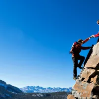 Kuvassa toinen vuorikiipeilijä auttaa toista nousemaan terävän vuorenhuipun päälle.