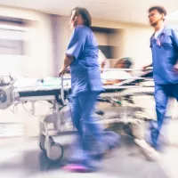 Kuvassa kaksi sairaanhoitajaa sinisissä työasuissa kiidättää potilasta potilasvuoteella sairaalan käytävällä oikealta vasemmalle.
