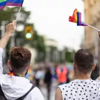 kaksi miestä kuvattuna selkäpuolelta, kun he marssivat Pride-liput käsissään kadulla.