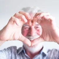 Harmaahiuksinen mies muodostaa käsillään sydämen kasvojensa eteen.