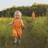 Lapsi kesäisellä niityllä, äiti kävelee kauempana taustalla.