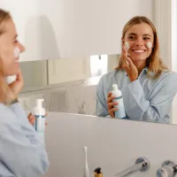 Nainen levittää voidetta kasvoilleen kylpyhuoneessa ja katsoo itseään samalla peilistä hymyillen.