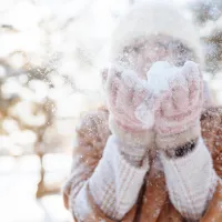 Talvisessa maisemassa henkilö pitelee käsissään lunta ja puhaltaa sen ilmaan, lumi pöllyää kauniisti auringonvalossa.