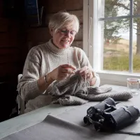 Tuovi Anttila neuloo mökillä pöydän ääressä ikkunan edessä.